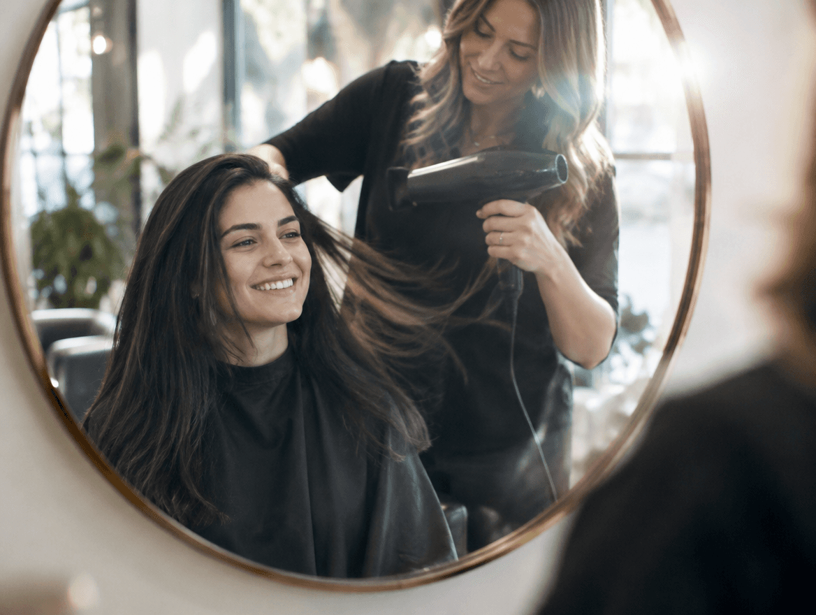 A stylist styling a smiling client's hair in a bright salon